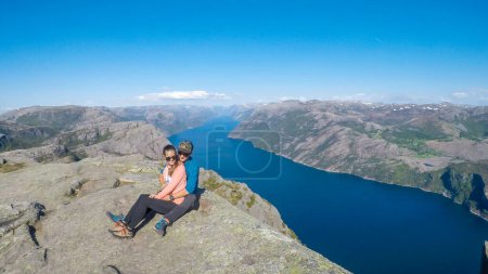 Téléchargez les photos : Un couple assis au bord d'une falaise escarpée de Preikestolen, avec vue sur Lysefjorden. Le fjord va loin à l'intérieur. Le couple profite de la vue, se sentant libre et heureux. Grande réalisation - en image libre de droit