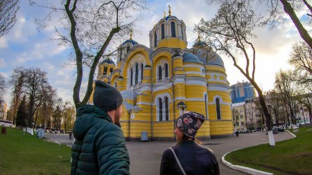 Téléchargez les photos : A couple taking a selfie in front of St Volodymyr's Cathedral. The cathedral is painted yellow, with blue and golden-domed rooftop. Overcast and cold day. Few trees on the side of the cathedral. - en image libre de droit