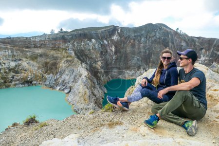 Foto de Pareja sentada en el borde del volcán y observando los lagos del cráter volcánico Kelimutu en Moni, Flores, Indonesia. Son relajados y tranquilos, disfrutando de la vista sobre el lago que brilla con muchos tonos de turquesa - Imagen libre de derechos