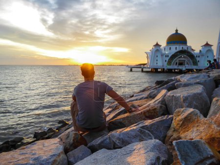 Un hombre sentado frente a la Mezquita del Estrecho de Malaca, Malasia. Es Patrimonio de la Humanidad. Tierna captura durante el atardecer, el sol se pone en el mar. Viajero individual.