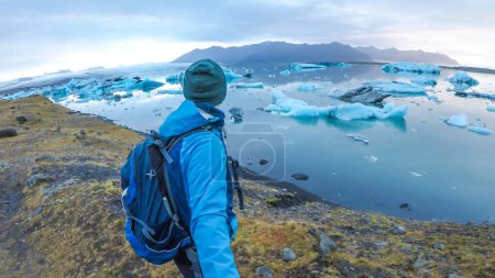 Foto de Un joven con una chaqueta azul y una mochila de senderismo contempla la laguna glaciar desde una orilla de hierba. Se está tomando una selfie con un palo. Bergs de hielo a la deriva en la laguna. Tranquilo y tranquilo. - Imagen libre de derechos