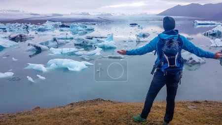 Foto de Un joven de pie en la orilla de una laguna glaciar, extiende sus manos. Bergs de hielo a la deriva hacia el mar. El calentamiento global derrite el paisaje helado. Encogimiento de glaciares. Admirando el fenómeno - Imagen libre de derechos