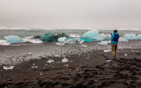 Foto de Un joven vistiendo una chaqueta azul se para en la playa de arena negra, sosteniendo su cámara de acción y filmando el mar agitado lavando los bergs de hielo a la orilla. Tonos azules de hielo. Efectos del calentamiento global. - Imagen libre de derechos