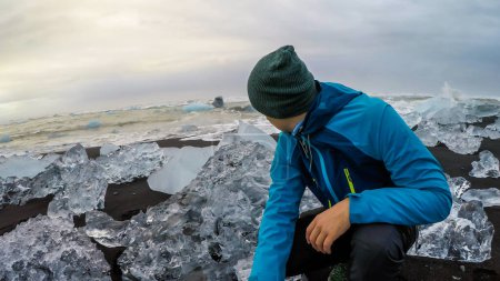 Foto de Hombre joven con chaqueta azul se sienta en una playa de diamantes, playa de arena negra y se toma una selfie mientras mira el mar. Disfruta de la belleza de este lugar. Mar áspero lanza más bergs de hielo en la orilla - Imagen libre de derechos