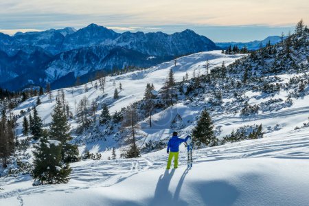 Foto de Cloudy morning in Alps, Nassfeld, Austria. Young skier walks on the powder snow with his skies. Mountain slopes covered with snow. Sharp edges of the Alps. Pine trees covered with snow on the slopes. - Imagen libre de derechos