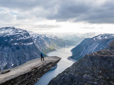 Téléchargez les photos : A man wearing blue jacket stands at the hanging rock formation, Trolltunga with a view on Ringedalsvatnet lake, Norway. Slopes of the mountains are partially covered with snow. Freedom and happiness - en image libre de droit