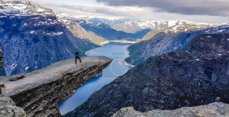 Foto de A man wearing blue jacket stands at the hanging rock formation, Trolltunga with a view on Ringedalsvatnet lake, Norway. Slopes of the mountains are partially covered with snow. Freedom and happiness - Imagen libre de derechos