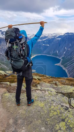 Foto de A young man wearing blue jacket and huge hiking backpack stands on a rock, holding a stick above his head. He admires the view in front of him - tall snow-capped mountains and navy blue lake. - Imagen libre de derechos