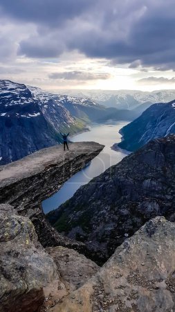 Téléchargez les photos : A man wearing blue jacket stands at the hanging rock formation, Trolltunga with a view on Ringedalsvatnet lake, Norway. Slopes of the mountains are partially covered with snow. Freedom and happiness - en image libre de droit