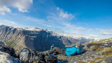 Foto de A man in blue jacket having breakfast in the tall mountains with a view on a fjord-like lake. He boils water in portable stove for morning coffee. Clear and sunny weather. Camping in the wilderness - Imagen libre de derechos
