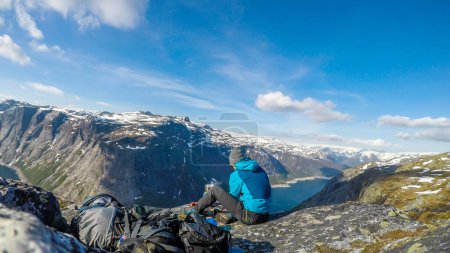 Foto de A man in blue jacket having breakfast in the tall mountains with a view on a fjord-like lake. He boils water in portable stove for morning coffee. Clear and sunny weather. Camping in the wilderness - Imagen libre de derechos