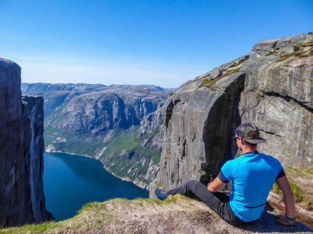 Foto de A young man sitting at the edge of a steep mountain. His legs are hanging down. Overcoming the fear of heights. In front of her stunning Lysefjorden shimmering with many shades of blue and green - Imagen libre de derechos