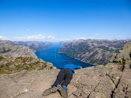 Téléchargez les photos : Un homme portant un T-shirt bleu penché sur le bord d'une falaise escarpée de Preikestolen, Pulpit Rock, avec une vue sur Lysefjorden. L'homme essaie de surmonter sa peur de la hauteur. Surmonter l'acrophobie - en image libre de droit