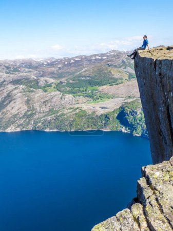 Téléchargez les photos : Un homme portant une chemise bleue est assis au bord d'une falaise escarpée de Preikestolen, les jambes pendantes. Une vue sur Lysefjorden. Le fjord va loin à l'intérieur. L'homme aime la vue, se sent libre et heureux. - en image libre de droit