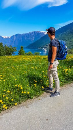 Téléchargez les photos : A young man with a hiking backpack looking at the picturesque view on port located at the Sognefjord, Norway. Few ships parked in the port. In front a meadow covered with yellow flowers. - en image libre de droit