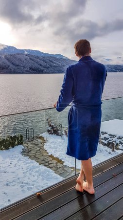 Photo for A man in a blue bathrobe standing on the balcony of a SPA in Austria in winter.  He is enjoying the view on Millstaetter lake and mountains in front of her. He is feeling relaxed and chilled. - Royalty Free Image