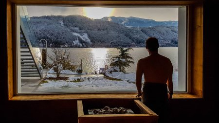 Photo for Man standing next to a window in an alpine sauna with the view on the lake and the mountains. Under the window there is a furnace, warming the stones. Mountains covered with snow Relaxation and chill - Royalty Free Image