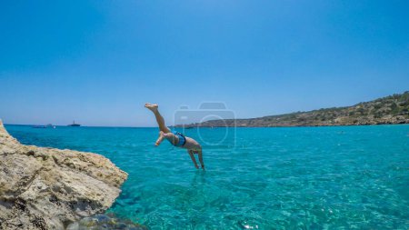 Photo for A young athletic-build man jumping into the sea in Cape Greco, Cyprus. Water is crystal clear, shimmering with many shades of blue. Jump of joy. Man is wearing blue swimsuit. - Royalty Free Image