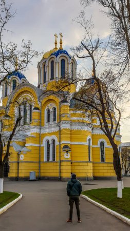 Téléchargez les photos : Un jeune homme vêtu d'une épaisse veste se tient devant la cathédrale Saint-Volodymyr. La cathédrale est peinte en jaune, avec un toit en dôme bleu et doré. Jour couvert et froid. - en image libre de droit