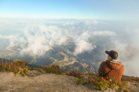 Foto de Un joven sentado en la cima del volcán Inerie en Bajawa, Flores, Indonesia. Está disfrutando de la hermosa vista de la isla volcánica. Hay algunas nubes debajo de él. Descubriendo mientras viaja. - Imagen libre de derechos