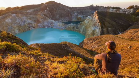 Foto de Un hombre observando el amanecer sobre los lagos del cráter volcánico Kelimutu en Moni, Flores, Indonesia. El horizonte es naranja, el sol sale lentamente. El hombre está relajado y tranquilo, disfrutando de la vista sobre el lago turquesa - Imagen libre de derechos