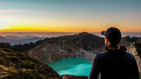 Foto de Un hombre observando el amanecer sobre los lagos del cráter volcánico Kelimutu en Moni, Flores, Indonesia. El horizonte es naranja, el sol sale lentamente. El hombre está relajado y tranquilo, disfrutando de la vista sobre el lago turquesa - Imagen libre de derechos