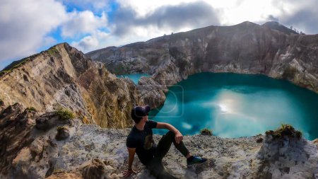 Foto de Hombre sentado en el borde del volcán y observando los lagos del cráter volcánico Kelimutu en Moni, Flores, Indonesia. El hombre está relajado y tranquilo, disfrutando de la vista sobre el lago que brilla con muchos tonos de turquesa - Imagen libre de derechos