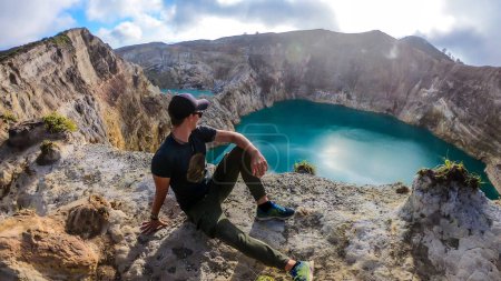 Foto de Hombre sentado en el borde del volcán y observando los lagos del cráter volcánico Kelimutu en Moni, Flores, Indonesia. El hombre está relajado y tranquilo, disfrutando de la vista sobre el lago que brilla con muchos tonos de turquesa - Imagen libre de derechos
