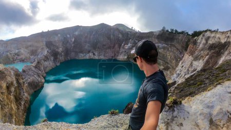 Foto de Hombre parado en el borde del volcán y observando los lagos del cráter volcánico Kelimutu en Moni, Flores, Indonesia. El hombre está relajado y tranquilo, disfrutando de la vista sobre el lago que brilla con muchos tonos de turquesa - Imagen libre de derechos