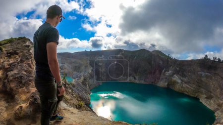 Foto de Hombre parado en el borde del volcán y observando los lagos del cráter volcánico Kelimutu en Moni, Flores, Indonesia. El hombre está relajado y tranquilo, disfrutando de la vista sobre el lago que brilla con muchos tonos de turquesa - Imagen libre de derechos