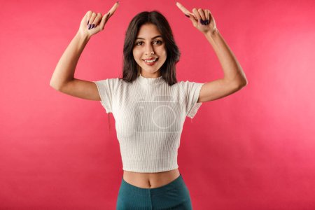 Foto de Happy young woman wearing white ribbed crop isolated over red background pointing fingers up at copy space. New product sharing, raise hands pointing fingers up smiling broadly. - Imagen libre de derechos