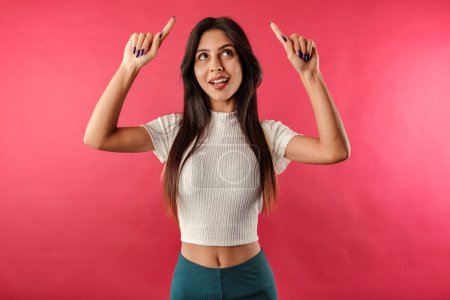 Foto de Happy brunette woman wearing casual top isolated over red background points upwards with excited looks with index fingers. Looks in amazement as indicates at something upwards. - Imagen libre de derechos