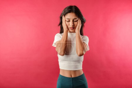 Foto de Portrait of young brunette woman wearing white ribbed crop isolated over red background holding hands on either side of head looks very tired and bored. Overwhelmed, wants to relax. - Imagen libre de derechos