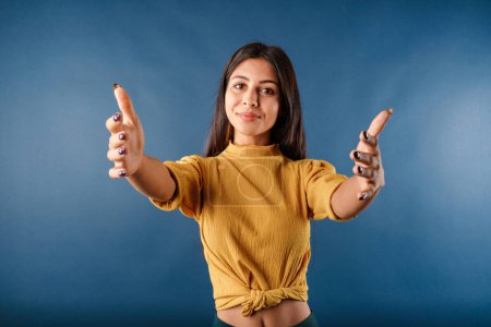Photo for Beautiful brunette woman wearing mustard yellow t-shirt isolated over blue background looking at the camera smiling with open arms for hug. Cheerful expression embracing happiness. Got some good news. - Royalty Free Image