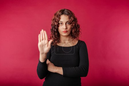 Portrait of young redhead woman wearing black ribbed dress isolated over red background doing stop sing with palm of the hand. Warning expression with negative and serious gesture on the face.