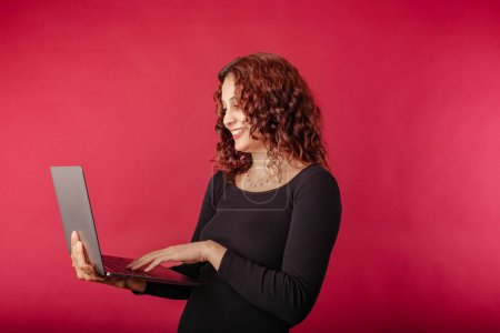 Redhead woman standing isolated over red background holding a laptop with a confident expression. Typing on laptop keyboard. Creating web design or digital work. Looking at the computer screen.