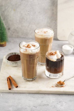 Photo for Three tall glasses with warm coffee drink with cinnamon, whipped milk foam and caramel on a marble board and christmas ornaments and decoration - Royalty Free Image