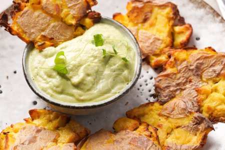 Trendy comfort snack crispy roasted crushed potatoes on a round plate with creamy avocado and garlic dip on a beige colored table cloth, close up