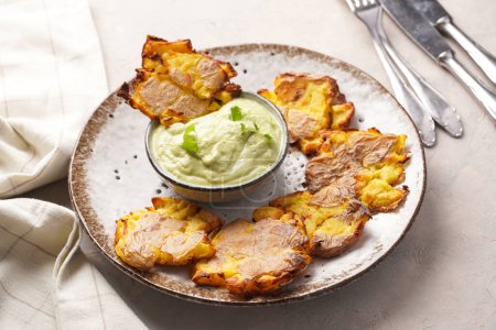 Trendy comfort snack crispy roasted crushed potatoes on a round plate with creamy avocado and garlic dip on a beige colored table cloth