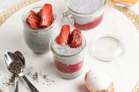 Breakfast idea - glass mason jars with chia seeds pudding, frozen strawberries and red raspberry smoothie on round marble board with easter decoration: golden eggs and white rabbits