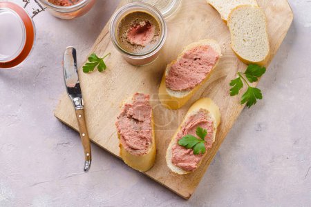 Homemade chicken liver pate on fresh french white wheat baguette slices on wooden board, glass mason jars with cooked liverwurst, top view