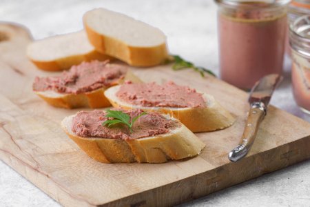 Homemade chicken liver pate on fresh french white wheat baguette slices on wooden plate, glass mason jars with cooked liverwurst