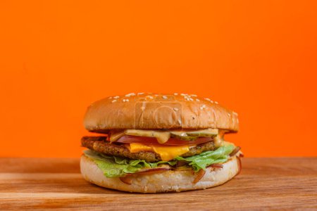 Delicious cheeseburger with lettuce, tomato, cheese, and bacon on a wooden surface. Studio shot with orange background. Fast food and indulgence concept. Design for menu, poster, and banner.
