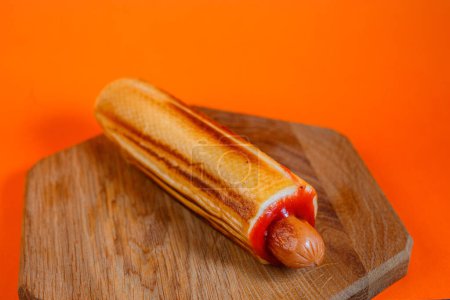 Grilled hot dog with ketchup on a toasted bun on a wooden cutting board. Studio shot with orange background. Classic American fast food concept. Design for menu, poster, and banner.