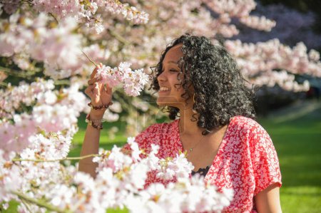 Téléchargez les photos : Femme métisse sentant l'odeur des fleurs de cerisier au printemps dans un parc - en image libre de droit