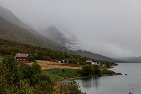 Foto de Casas a los pies de montañas brumosas. Granja roja y campos. Paisaje noruega. - Imagen libre de derechos