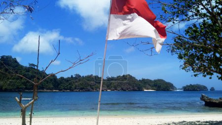 Foto de Bandera de Indonesia ondeando en una playa durante el día soleado bajo el cielo azul claro. Océano e isla en el fondo. - Imagen libre de derechos