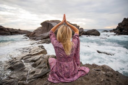 Téléchargez les photos : Fille médite assis sur les rochers regardant l'océan et elle fait du yoga, concept de spiritualité - en image libre de droit