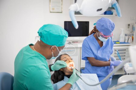 Foto de Un dentista africano comprueba la salud bucal de su paciente mientras su asistente prepara las herramientas de trabajo. - Imagen libre de derechos