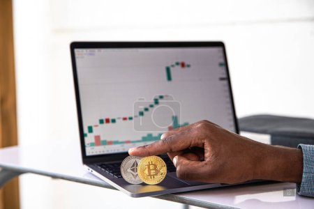 A cryptocurrency trader's hand holding a Bitcoin and Ethereum coin with the laptop with the market graph behind it.